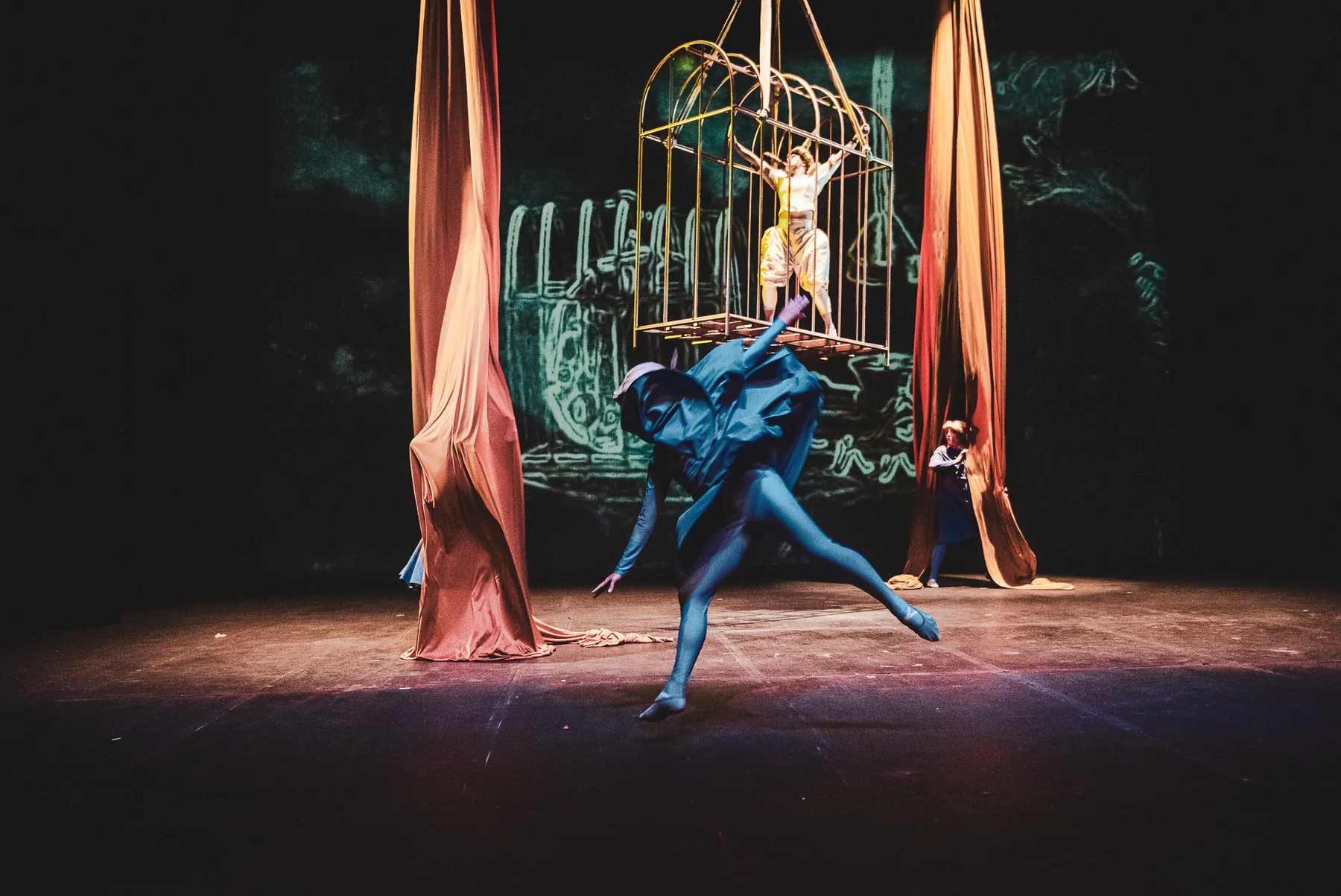 Un personaje de azul danza frente a proyecciones y aparatos suspendidos.